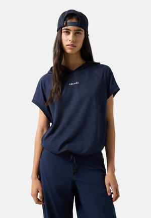 Młoda kobieta ubrana w granatową bluzę z kapturem z logo "FIRE+ICE", pasujące spodnie oraz granatową czapkę z daszkiem noszoną do tyłu, stoi na jednolitym tle.