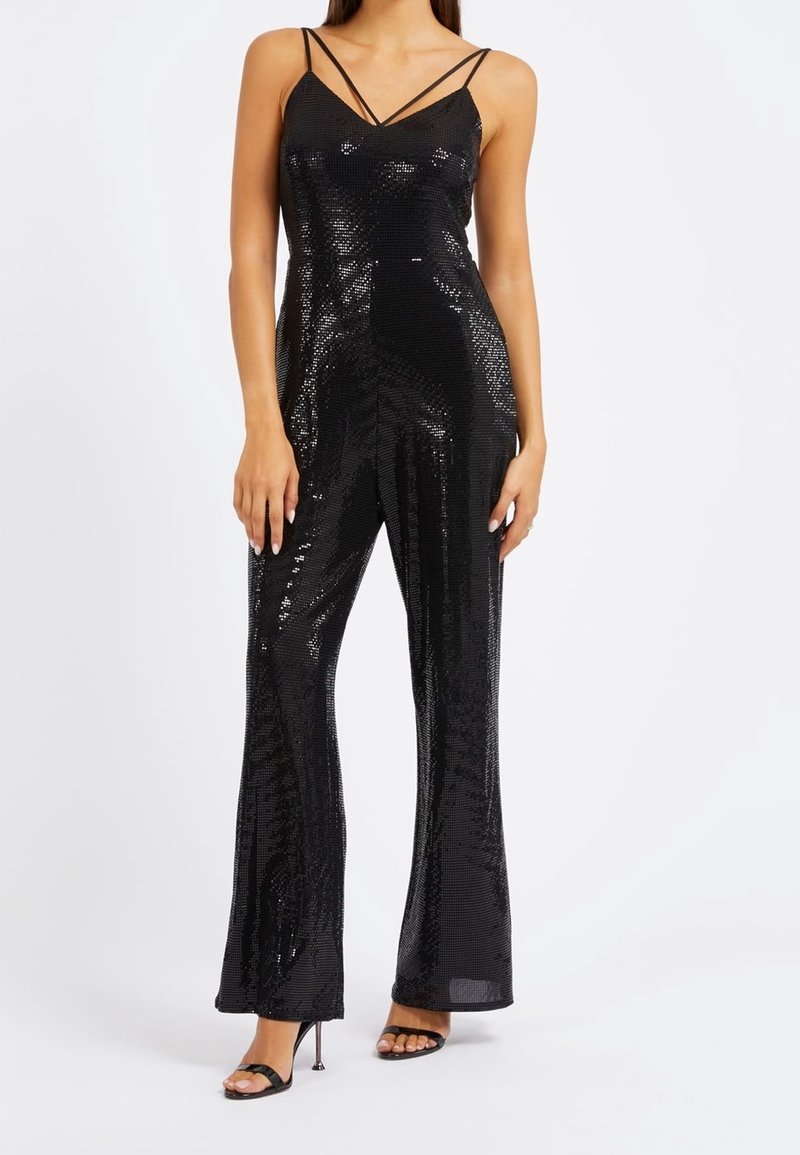 Femme portant une combinaison noire à sequins avec double bretelles fines et pantalon à jambes larges, associée à des sandales noires à talons hauts.