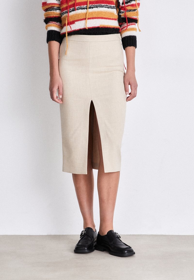 Personne portant une jupe beige mi-longue fendue devant, un pull rayé multicolore et des mocassins en cuir noir, debout sur un fond blanc.