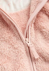 Veste zippée en polaire rose avec un extérieur doux et texturé, dotée d'une fermeture éclair assortie avec une languette, révélant un intérieur blanc avec un motif subtil.