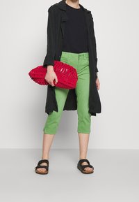 Svart lång kappa, svart t-shirt, gröna cropped-byxor och svarta sandaler. Håller en röd, strukturerad vävd clutch-väska.