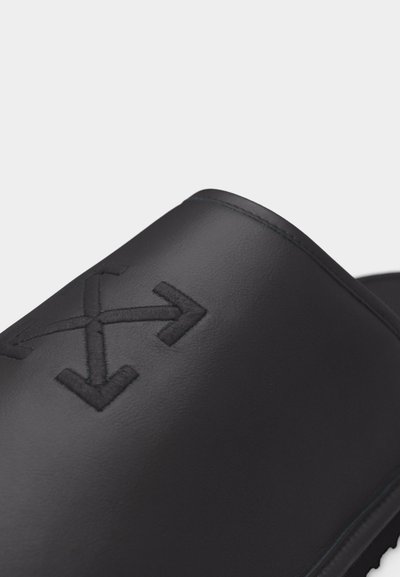 Μαύρη σανδάλι με σαγιονάρα με ανάγλυφο λογότυπο που εμφανίζει τέσσερα βέλη που δείχνουν προς τα έξω από ένα κεντρικό σχήμα X στο λουρί.