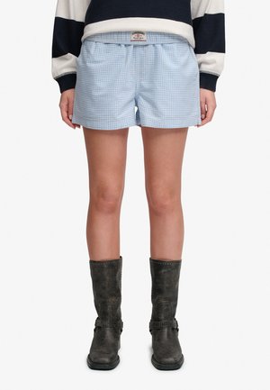 Person iført lyseblå og hvide ternede shorts med elastisk talje, sorte støvler til midt på læggen med stropper og en sort-hvid stribet langærmet top.