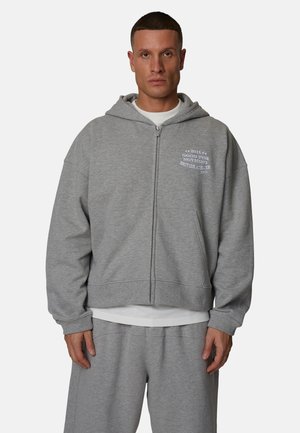 Felpa grigia con zip e vestibilità ampia, dotata di tasca frontale, cappuccio con coulisse e testo grafico sul petto. Materiale morbido in pile.