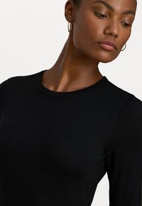 Top de punto negro de manga larga con escote redondo, que presenta una textura suave y detalles de costura minimalistas. Acentuado con pendientes de aro.