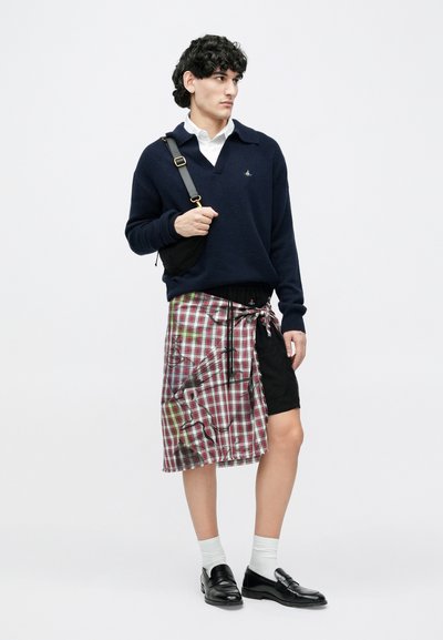 Vyras su garbanotais plaukais, vilkintis tamsiai mėlyną megztinį ant baltos marškinės, juodai raudoną languotą sijoną, baltas kojines, juodus batus ir nešiojantis juodą per petį kabočią rankinę.