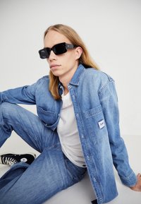 Denimowa koszula i dżinsy w jasnoniebieskim kolorze, z guzikowym zapięciem i kieszenią, zestawione z czarnymi, masywnymi okularami przeciwsłonecznymi oraz białą koszulką.