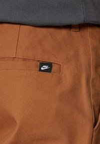 Brown cotton pants with a smooth texture and a back pocket featuring a black Nike logo tag. Gray fabric peeks from above.