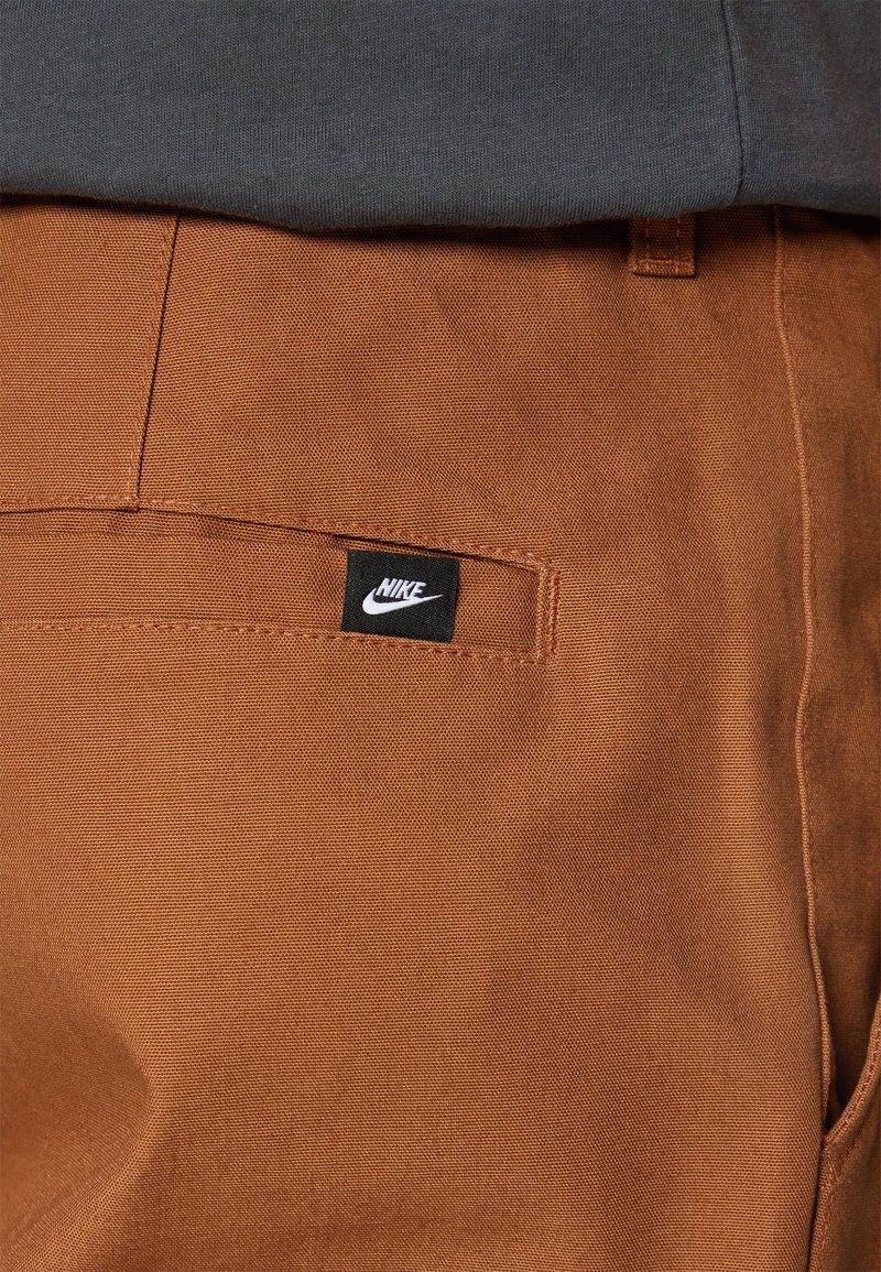 Pantalons en coton marron avec une texture lisse et une poche arrière arborant une étiquette noire avec le logo Nike. Un tissu gris apparaît par-dessus.