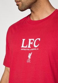 Κόκκινο βαμβακερό μπλουζάκι με άσπρο κείμενο «LFC» και «ANFIELD RD, LIVERPOOL, L4», με το έμβλημα τ�ης Liverpool FC και το «L.F.C.» από κάτω.