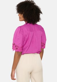 Blusa rosa con maniche a sbuffo e bordo in pizzo ai polsini. Il retro presenta un dettaglio plissé. Abbinata a pantaloni beige.