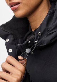 Persona che regola il cappuccio di una giacca nera con coulisse e fermo, concentrandosi sulla mano, la cerniera e la parte inferiore del volto.