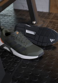 Paire de chaussures de sport vert foncé et noir avec semelles blanches reposant sur un sol de gym en caoutchouc noir texturé près d'un équipement métallique.