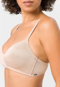 Beige Softcup-BH mit verstellbaren Trägern, nahtlosem Design und subtiler Sheen. Verfügt über einen kleinen metallischen Akzent an der Seite.