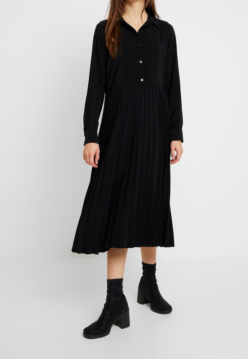 Robe noire à manches longues avec boutons sur le devant et jupe plissée, en tissu lisse. Assortie à des bottines noires, le tout sur un fond neutre.