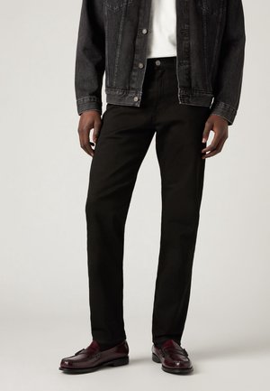 Homme portant un jean noir, une veste en denim foncé, une chemise blanche et des mocassins en cuir rouge foncé, se tenant devant un fond uni.