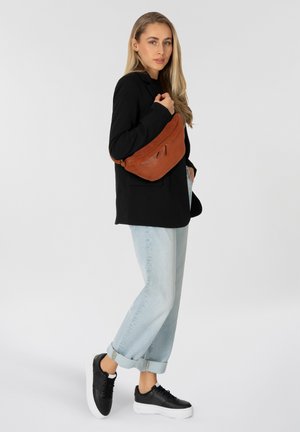 Borsa a tracolla in pelle marrone con chiusura a zip, forma arrotondata, cuciture pronunciate. Indossata con un blazer nero e jeans azzurri risvoltati.