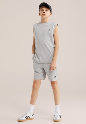 Garçon portant un débardeur gris et un short avec une casquette noire et des baskets blanches, debout devant un fond beige uni.