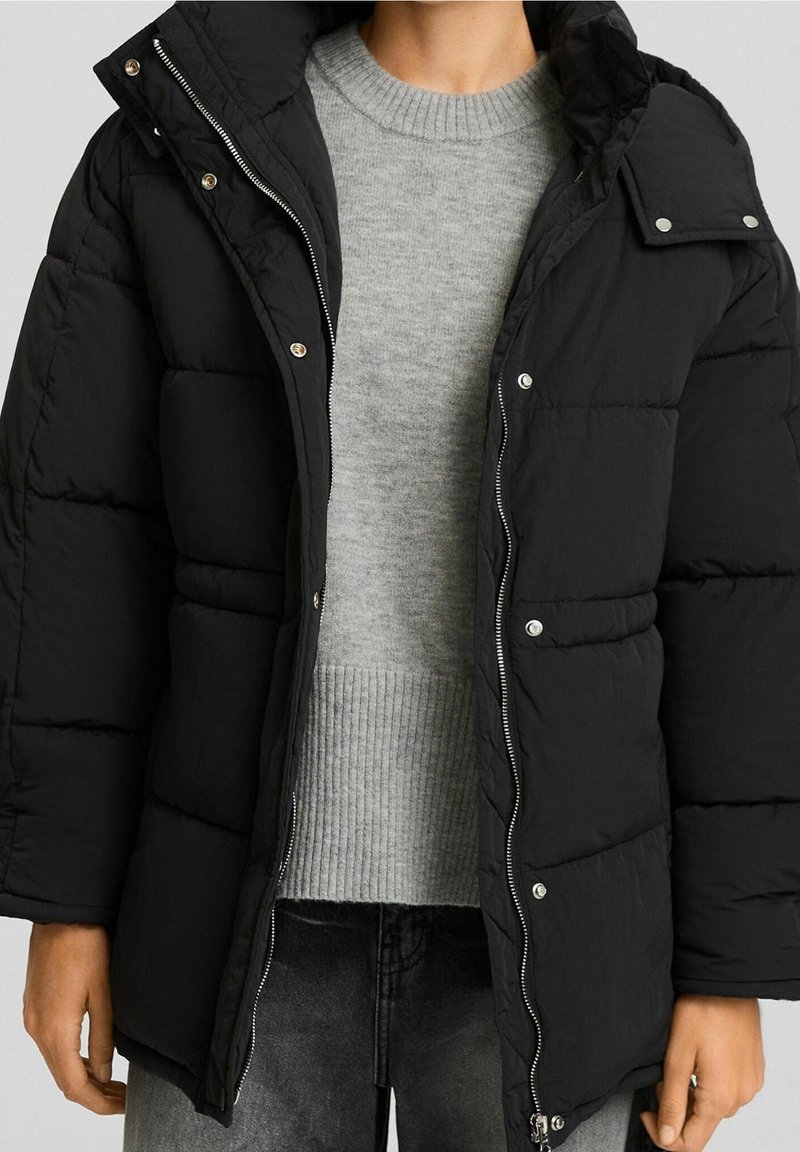 Svart puffjacka med dragkedja fram, tryckknappar och huva; tillverkad av ett slätt tyg med en quiltad design. Grå tröja under.