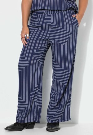 Personne portant un pantalon large bleu marine avec des rayures géométriques blanches, des chaussures noires et les mains dans les poches sur un fond clair uni.