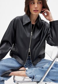 Juoda odinė trumpa striukė su dideliu apykakle ir dviem užtrauktukais; derinama su šviesiai mėlynomis džinsinėmis kelnėmis ir juodais aštriakraščiais bateliais.