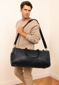Musta nahkainen duffel-laukku, jossa on kaksi kahvaa ja säädettävä olkahihna. Laukussa on sileä pinta ja kultaiset yksityiskohdat.