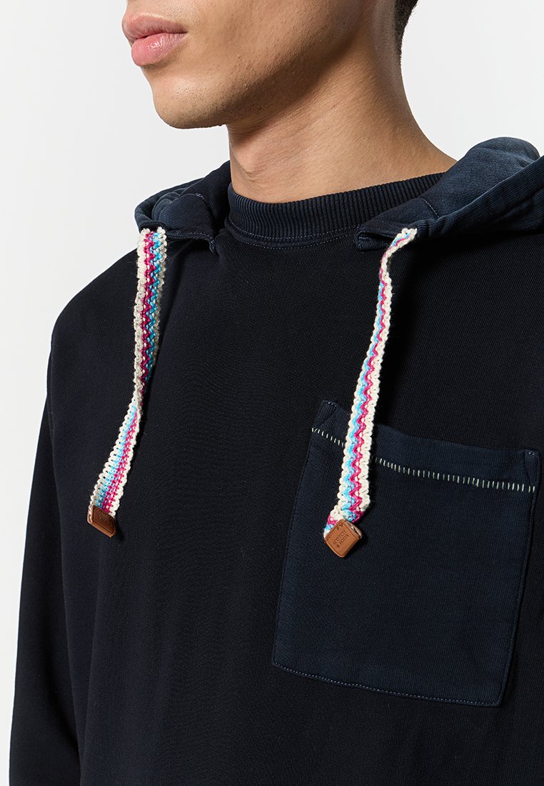 Ein Mann trägt einen dunkelblauen Kapuzenpullover mit mehrfarbigen, geflochtenen Kordeln und einer Brusttasche vor einem schlichten hellen Hintergrund.