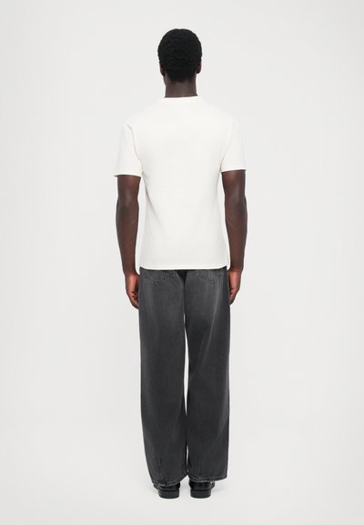 White short-sleeve t-shirt with a round neck, paired with loose-fitting dark gray jeans and black shoes, viewed from the back.