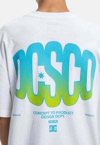 Weißes Baumwoll-T-Shirt mit auffälligem blauen und grünen Farbverlauf-Text "DCSC" sowie einem Stern und kleinerem Text darunter, der "CONCEPT TO PRODUCT" enthält.