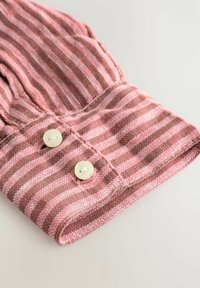 Poignet de chemise en tissu rayé rose et marron avec deux boutons blancs sur un fond clair.