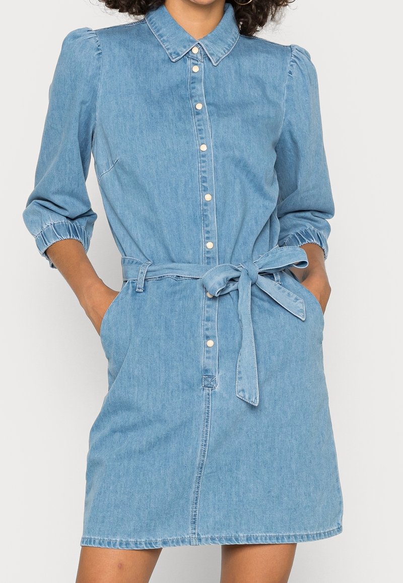 Robe en denim bleu clair avec un devant boutonné, un col classique, des manches élastiquées aux poignets et une ceinture en tissu à nouer à la taille. Comprend des poches latérales.