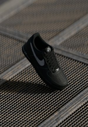 Zwarte Nike Air Force 1 sneaker met een glad leren bovenwerk, geperforeerde neus, grijze accenten en zwarte veters, zwevend boven een gestructureerd oppervlak.