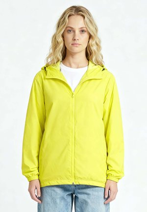 Blonde vrouw met een felgeel jack met rits en capuchon over een wit shirt en lichtblauwe jeans, staand voor een effen achtergrond.