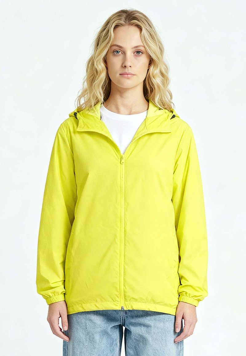 Femme blonde portant une veste zippée jaune vif avec capuche par-dessus un t-shirt blanc et un jean bleu clair, debout devant un fond uni.