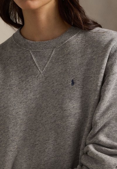Nainen, jolla on harmaa pyöreäkauluksinen collegepaita, jossa on rinnassa pieni tummanvärinen kirjailtu hevonen ja ratsastaja -logo.