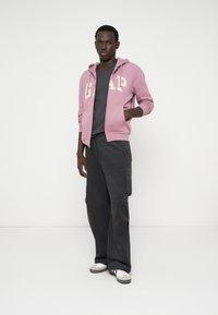 Ανοιχτό ροζ φούτερ με φερμουάρ και λογότυπο "GAP", φορεμένο πάνω από ένα σκούρο γκρι μπλουζάκι, συνδυασμένο με σκούρες φαρδιές παντελόνες και καλοκαιρινά αθλητικά παπούτσια.