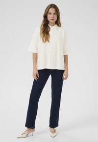 White, short-sleeve button-up shirt with a textured pattern; paired with dark blue jeans and pale, pointed-toe heels.
