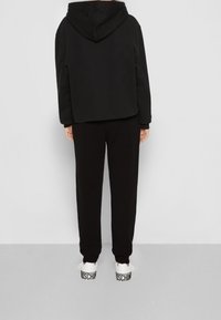 Schwarze Kapuzenjacke mit lockerer Passform, kombiniert mit passenden schwarzen Hosen und weißen Sneakers mit "MOSCHINO"-Branding. Durchgehend glatte Textur.