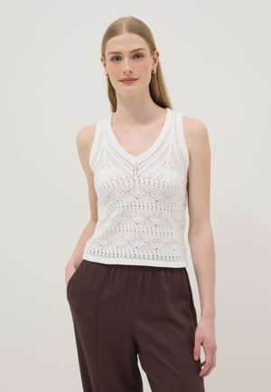 Jonge vrouw met steil haar, gouden oorringen in ringenvorm, een wit, mouwloos V-hals topje met patroon, en donkerbruine broek, staand met één hand in de zak.