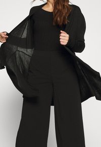 Černý dlouhý cardigan vyrobený z lehké, texturované pleteniny. Má otevřený přední díl, žebrované detaily a volný střih. Doplňuje ho černý top a kalhoty.