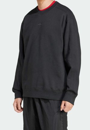 Osoba ubrana w luźną czarną bluzę z małym logo Adidas, czarne spodnie oraz czerwoną koszulkę z okrągłym dekoltem widoczną przy kołnierzu.
