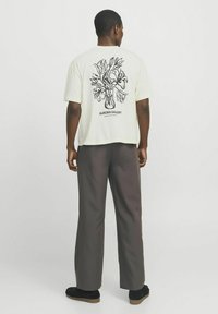 T-shirt couleur crème à manches courtes, présentant un graphique de vase floral noir au dos. Associé à un pantalon ample gris foncé.