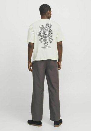 T-shirt couleur crème à manches courtes, présentant un graphique de vase floral noir au dos. Associé à un pantalon ample gris foncé.