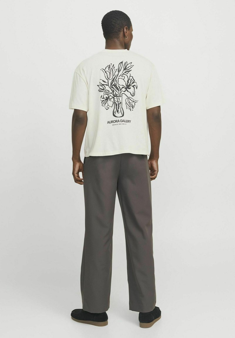 T-shirt couleur crème à manches courtes, présentant un graphique de vase floral noir au dos. Associé à un pantalon ample gris foncé.