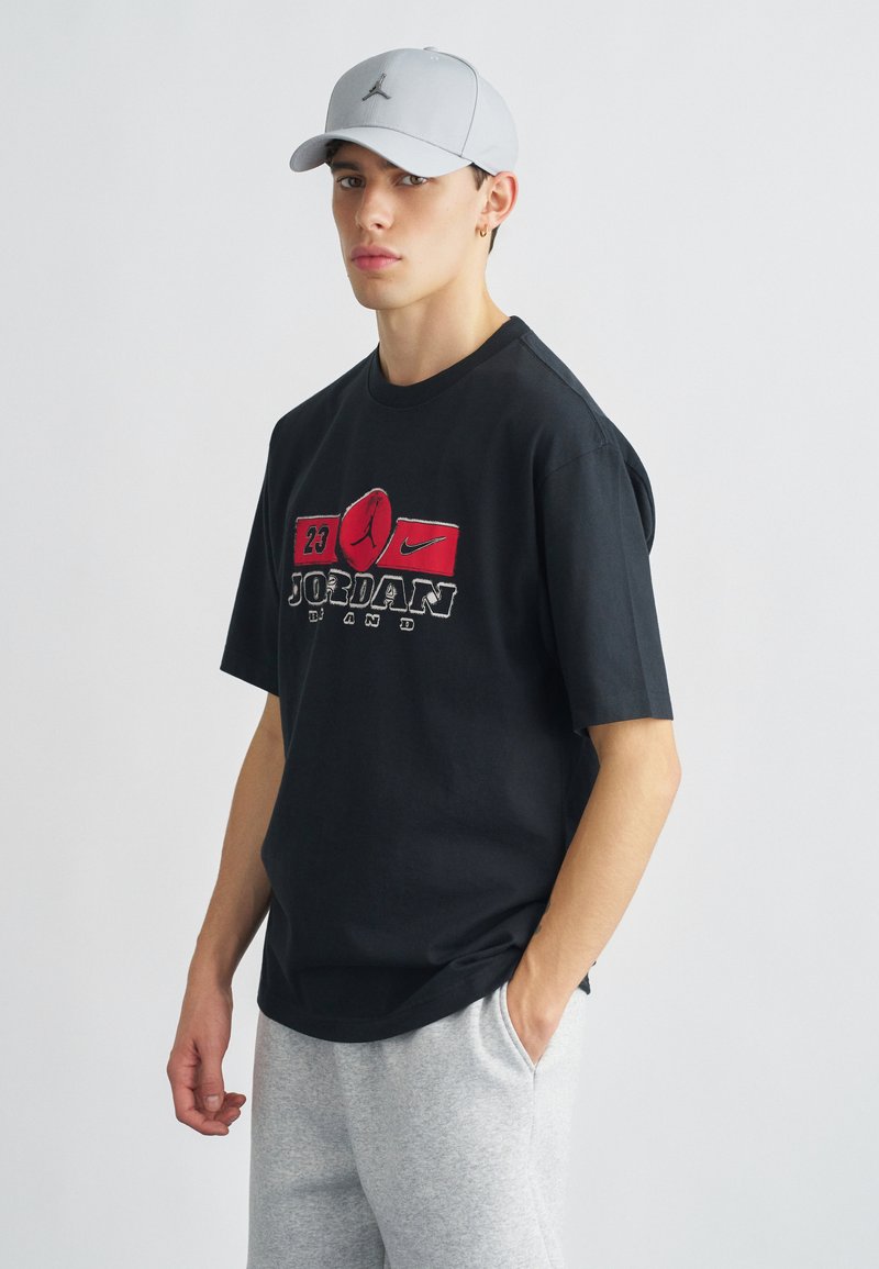 Μαύρο βαμβακερό μπλουζάκι με κόκκινο και λευκό λογότυπο Jordan. Συνδυασμένο με ανοιχτό γκρι παντελόνι φόρμας και ανοιχτό γκρι καπέλο baseball.