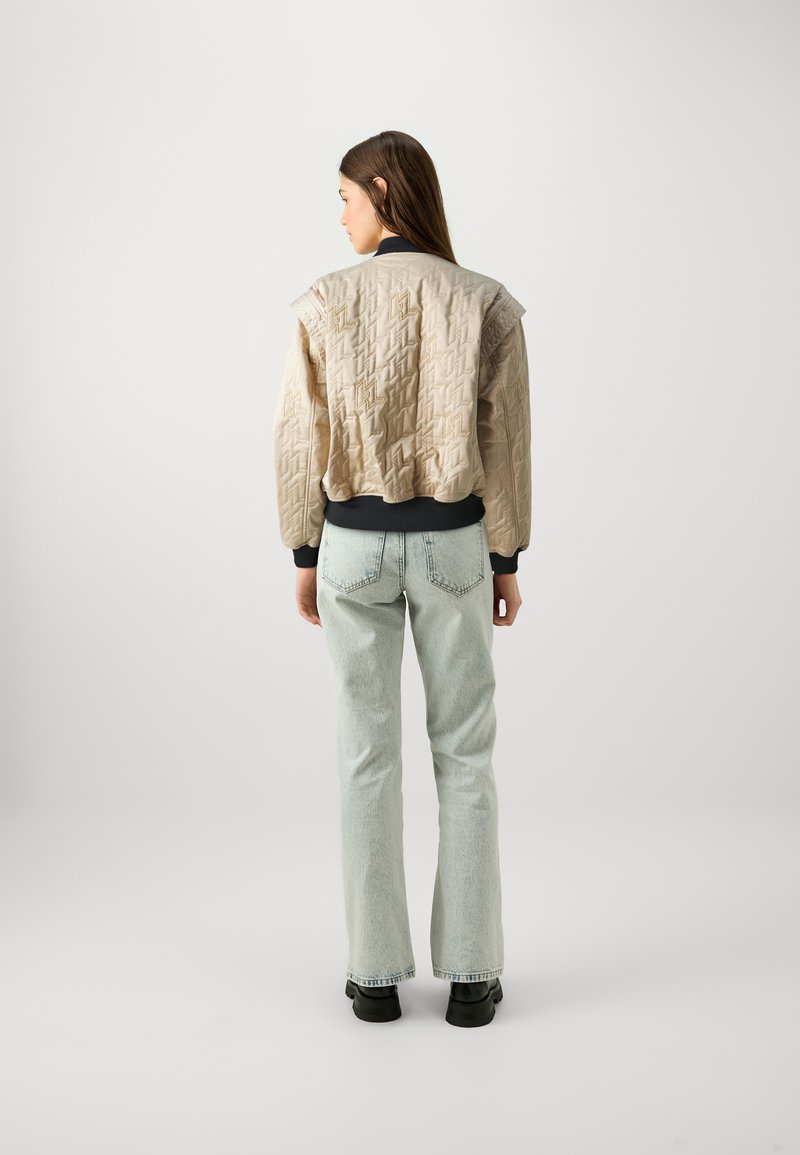 Beige, gesteppte Bomberjacke mit übergroßen Schultern, gerippten Bündchen und Saum; kombiniert mit hellblauen, ausgestellten Jeans und schwarzem Schuhwerk.