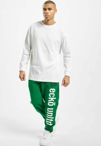 Hvid langærmet skjorte parret med grønne sweatpants med hvid "eckō united" tekst. Modellen bærer hvide sneakers. Enkel og afslappet design.