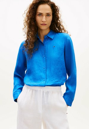 Kobieta o kręconych włosach, ubrana w jaskrawo niebieską koszulę na guziki i białe spodnie ze sznurkiem, stojąca z rękami w kieszeniach.