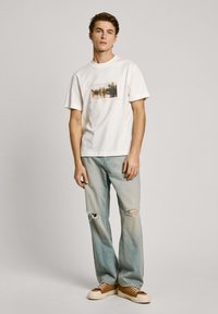 Balts T-krekls ar foto druku, kombinēts ar gaiši ziliem nolietotiem džinsiem un brūnām kurpēm. Ikdienišķs griezums, īsas piedurknes un minimāls dizains.