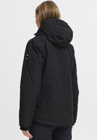 Veste imperméable noire avec capuche, présentant une texture lisse, des poignets élastiques et un logo sur la manches. Vue arrière montrée.